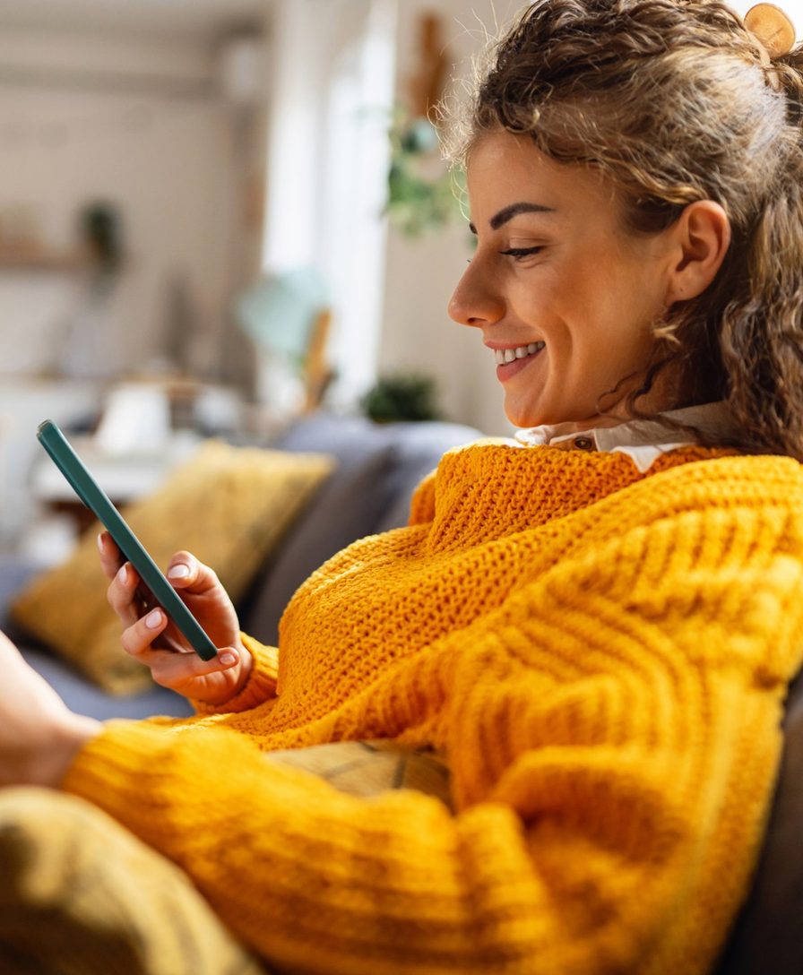 Imagem ilustrativa: mulher sorri usando o celular e segurando um boleto, sentada no sofá de casa