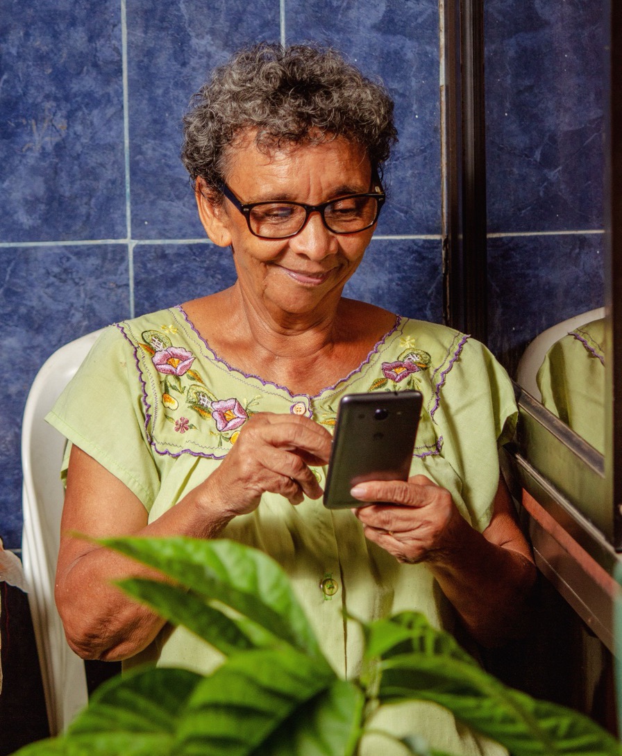 Imagem ilustrativa: mulher de óculos sorri olhando o celular, sentada em casa